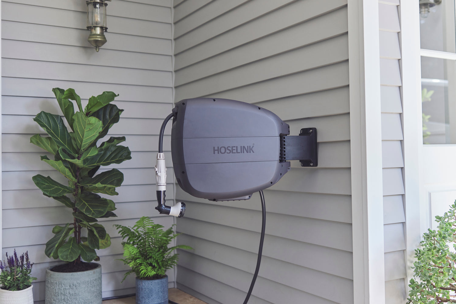 Charcoal Evolve Retractable Hose Reel mounted on on the side of a grey weatherboard house with some potted plants in the background