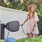 Red haired woman in pink dress and hat using a charcoal Evolve Retractable Hose Reel mounted on a black timber post to water a potted fern in the backyard