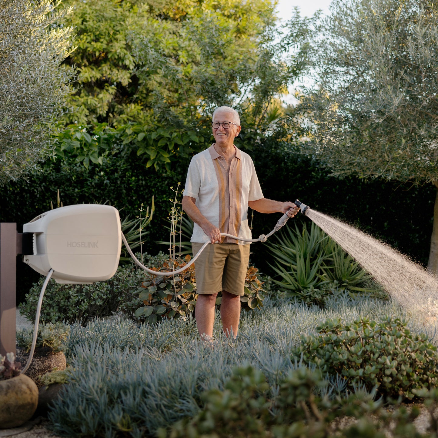 Older man in a linen shirt and brown shorts watering a succulent garden with a Beige Evolve Retractable Hose Reel mounted on a black timber post