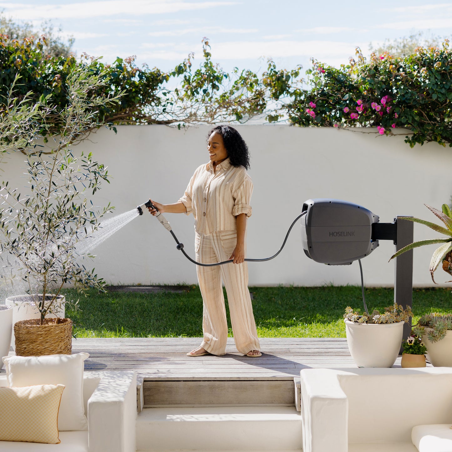 Woman standing in front of a white rendered wall on her wooden deck in the back yard watering a potted olive tree with an Evolve Charcoal Retractable Hose Reel