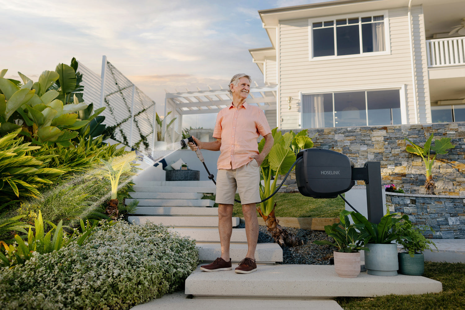 man in pink short and khaki shorts watering a garden bed with a charcoal Evolve Retractable Hose Reel mounted on a black timber post with a white house in the background