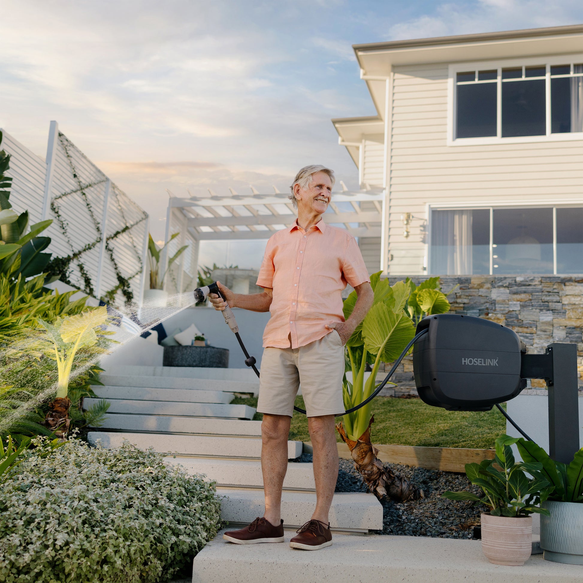 Older man in pink shirt and khaki shorts standing on white stone steps in front of a weather board house using a Charcoal Evolve Retractable Hose Reel to water plants