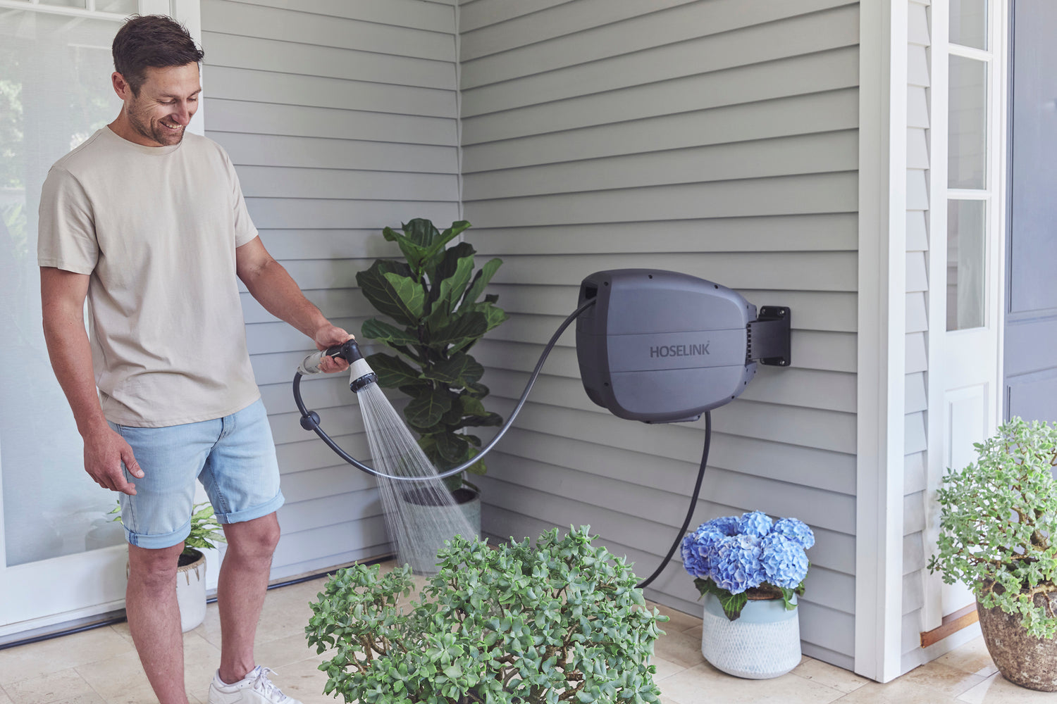 Ben Hayman in jean shorts and a beige shirt using a Charcoal Evolve Retractable Hose Reel mounted on  weatherboard home to water a potted plant