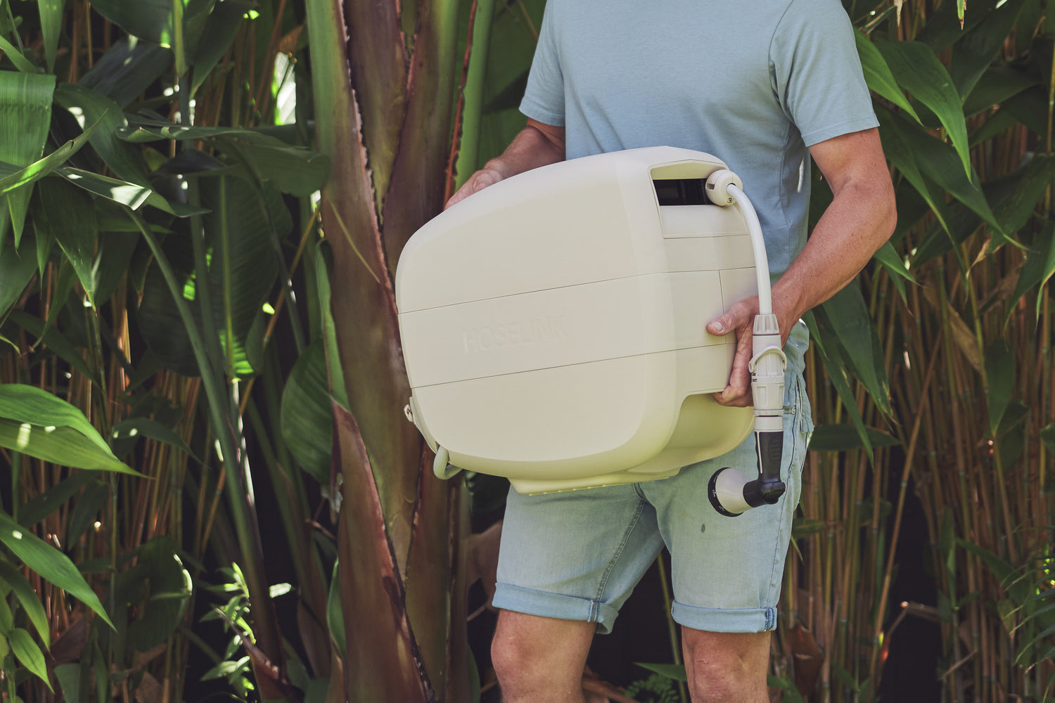 Man in light blue shirt and jean shorts carrying the Beige Evolve Retractable Hose Reel through the garden with bamboo in the background