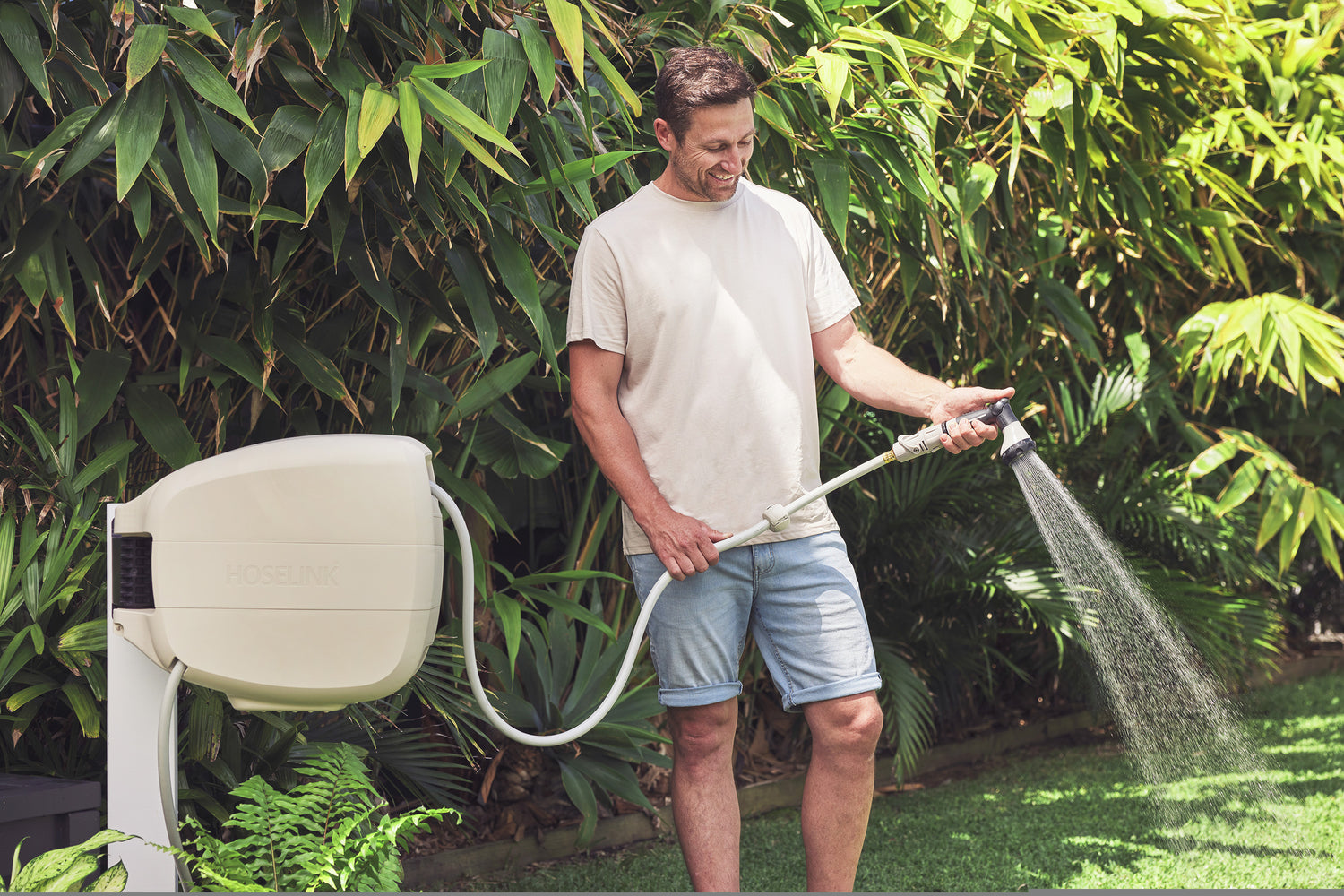 Ben Hayman in jean shorts and a beige shirt using a Charcoal Evolve Retractable Hose Reel mounted on  a white post to water the lawn