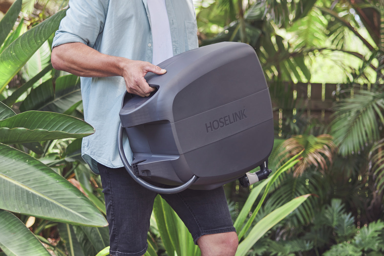 Man in light blue shirt and dark blue shorts carrying the Charcoal Evolve Retractable Hose Reel through the garden