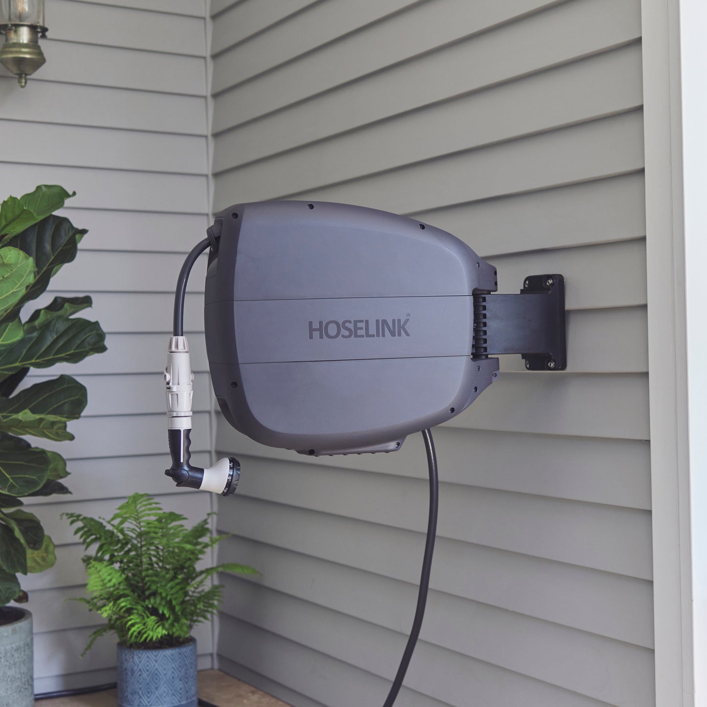 Evolve charcoal Retractable Hose Reel mounted on the corner of a grey weather board house with some potted ferns in the background