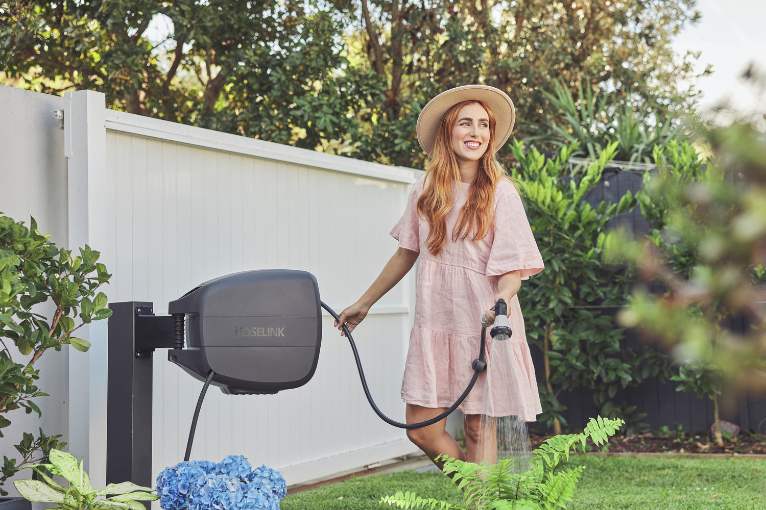 Evolve charcoal retractable hose reel mounted on a black post in a garden. Being used by a woman in a pink dress and hat to water a green fern