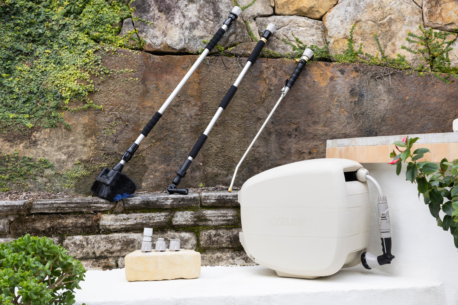 Beige Evolve Retractable Hose Reel sitting on a white step with an Extendable Cleaning Brush, Pivot Gutter Cleaner and Super Jet Washer Leaning against a stone wall and a warm grey Adapter Pack next to the reel