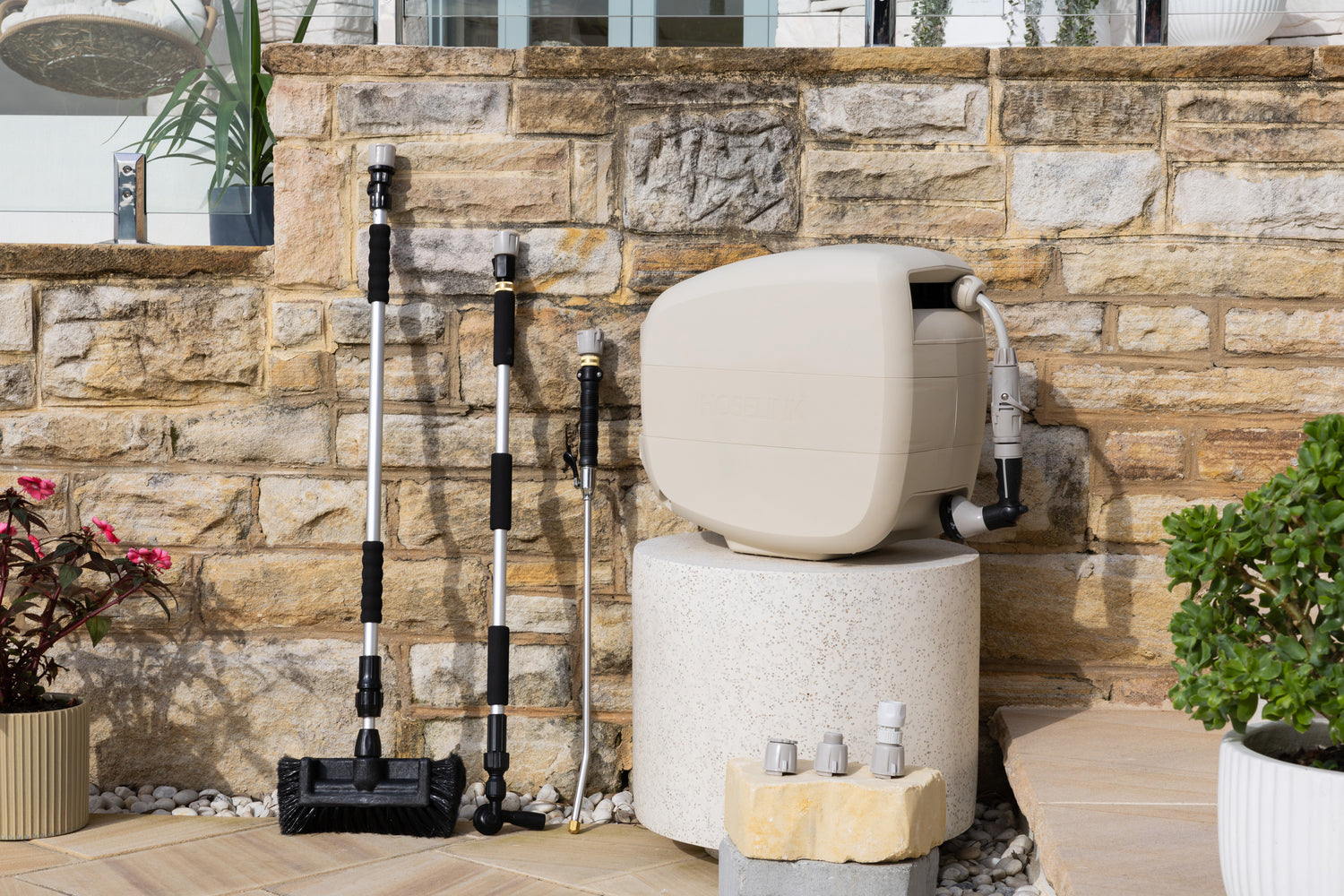 Evolve Beige Retractable Hose Reel on a white stone table with an adapter pack in front and Extendable Cleaning Brush, Super Jet Washer and Pivot Gutter Cleaner leaning on a sandstone wall next to the reel