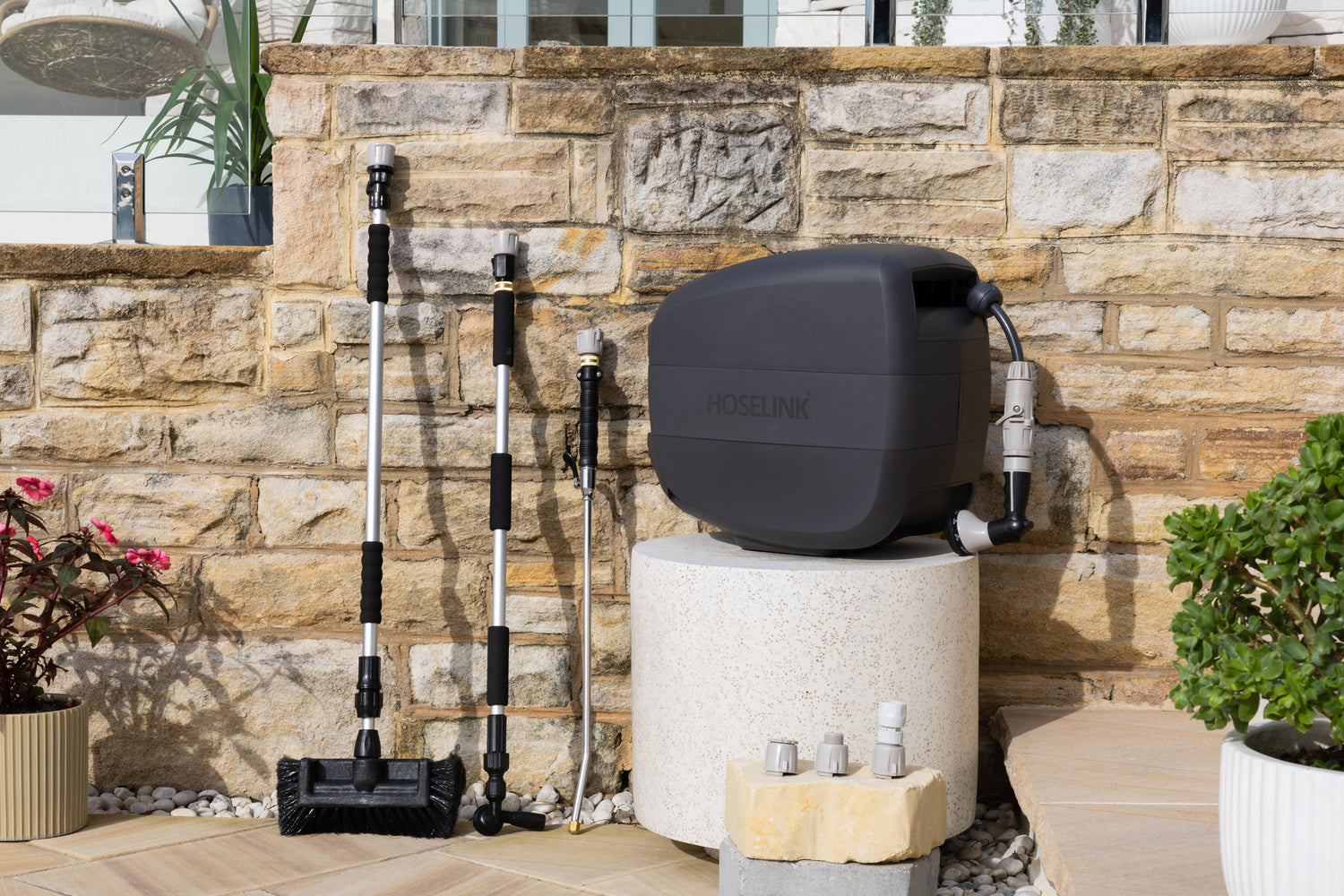 Charcoal Evolve Retractable Hose Reel on a white stone table with an Extendable Cleaning Brush, Super Jet Washer and Pivot Gutter Cleaner leaning on a sandstone wall next to the reel and an adapter pack in front of the reel 