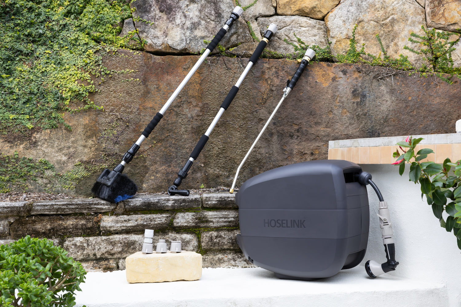 Evolve Charcoal Retractable Hose Reel sitting on a white step with an Adapter Pack next to it and an Extendable Cleaning Brush, Pivot Gutter Cleaner and Super Jet Washer Leaning against a stone wall in the background