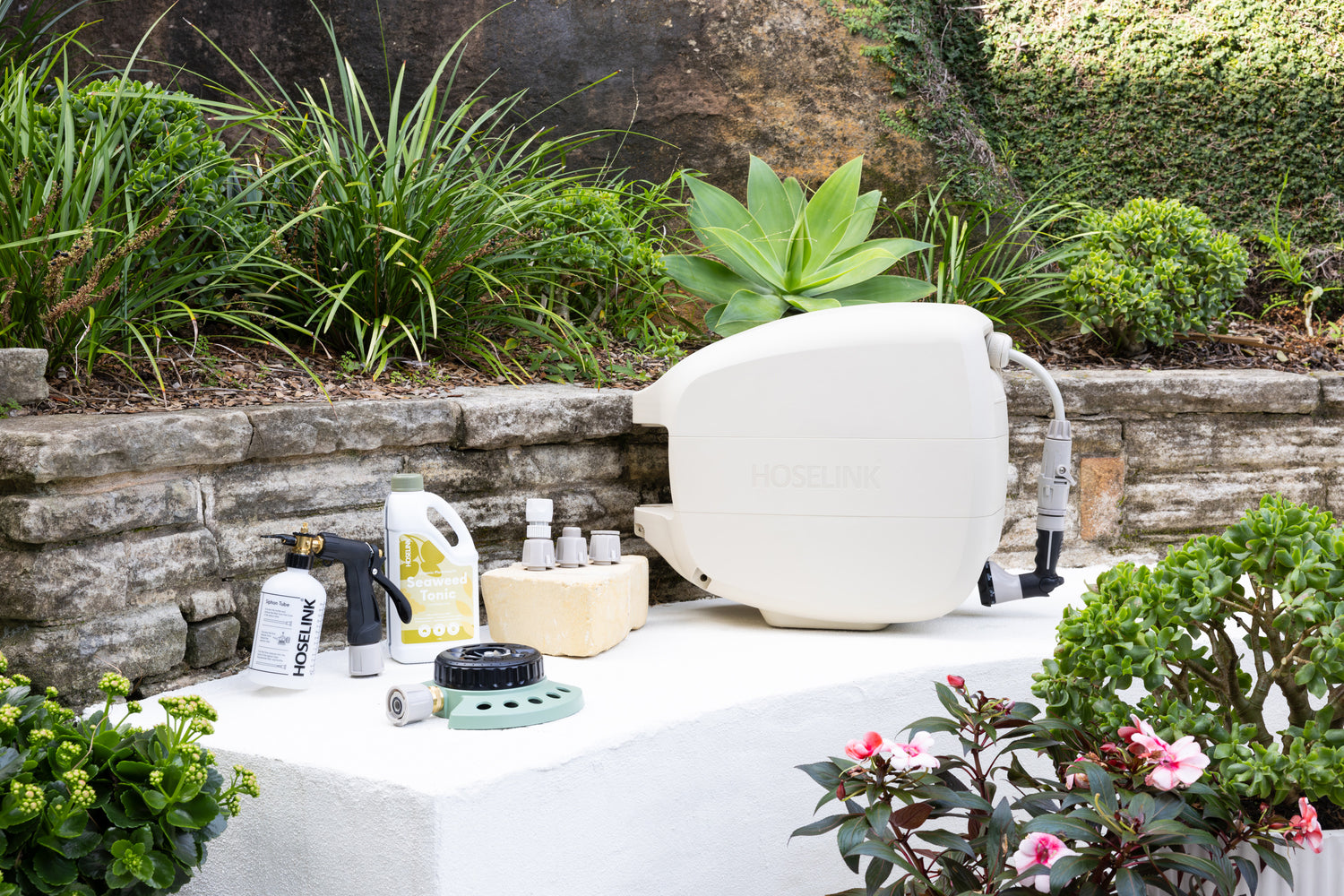 Evolve Beige Retractable Hose Reel sitting next to a Fertiliser Spray Mixer, Adapter Pack, 9-pattern Sprinkler and Seaweed Tonic on a white stone bench in front of a garden bed