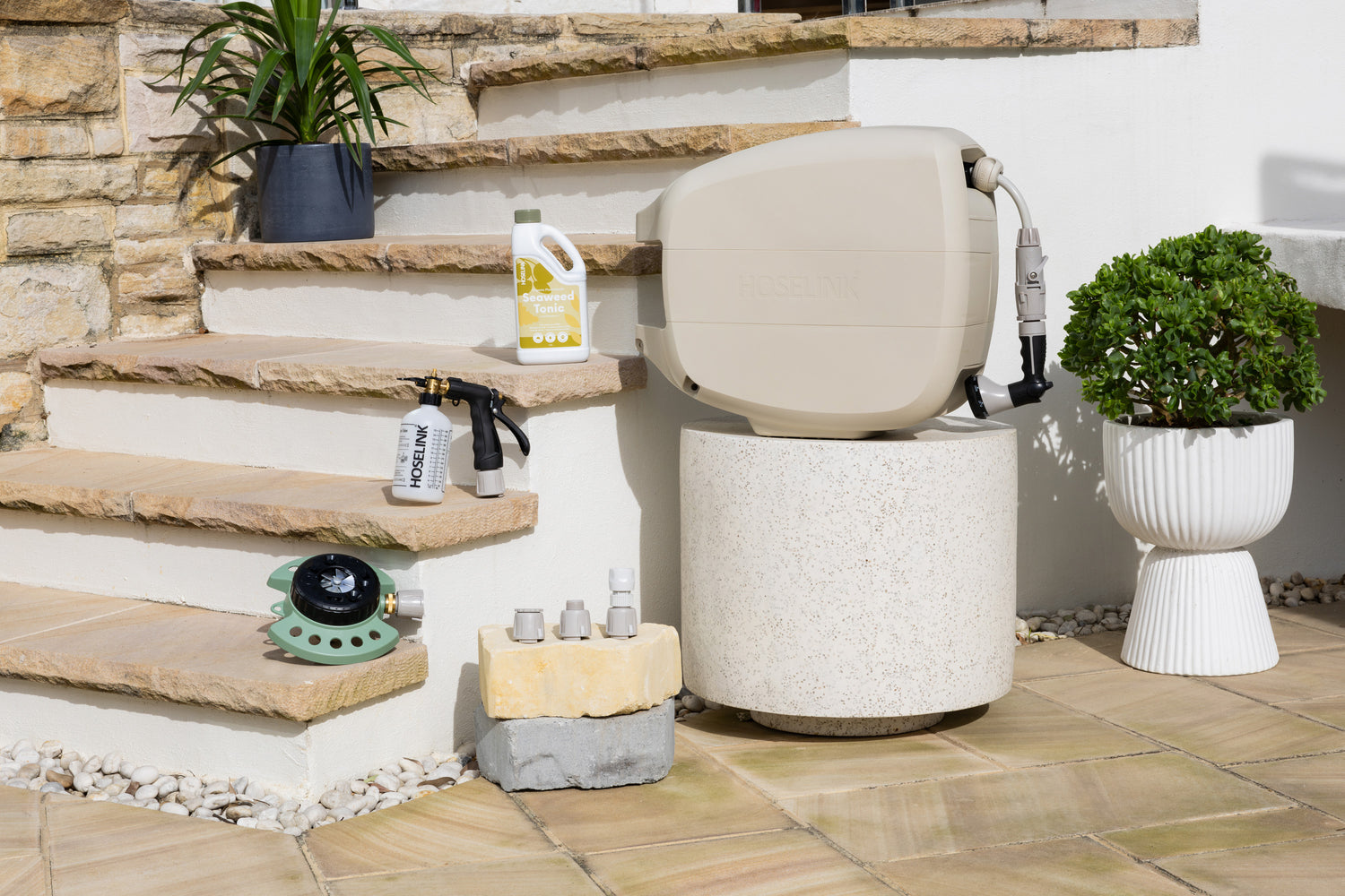 9-Pattern Sprinkler, Sea Weed Tonic Fertiliser Spray Mixer and Adapter Pack sitting on sandstone steps next to a white stone table with an Evolve Beige Retractable Hose Reel on it. 