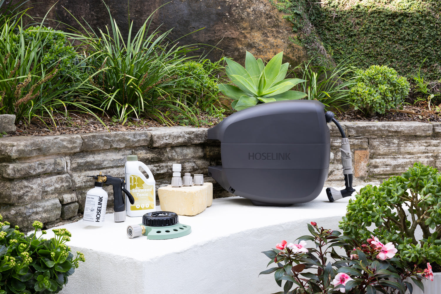 Charcoal Evolve Retractable Hose Reel sitting next to a Fertiliser Spray Mixer, Seaweed Tonic, Adapter Pack and 9-pattern Sprinkler on a white stone bench in front of a garden bed