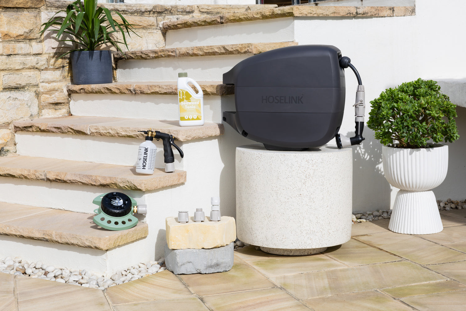 Fertiliser Spray Mixer, Sea Weed Tonic, 9-Pattern Sprinkler and Adapter Pack sitting on sandstone steps next to a white stone table with a Charcoal Evolve Retractable Hose Reel on it. 