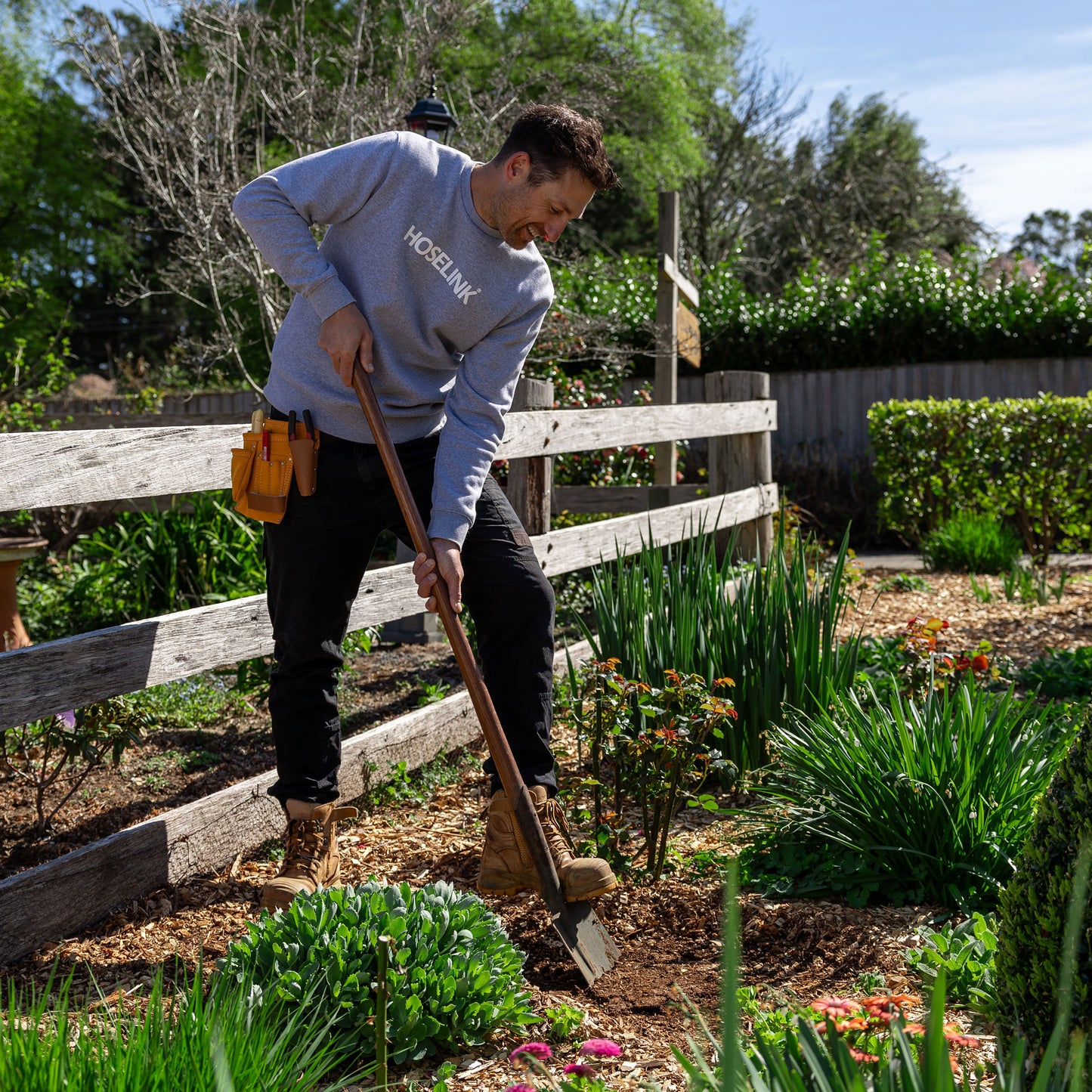 ben hayman using a shovel to dig a hole in a garden bed, next to a timber fence surrounded by green shrubs