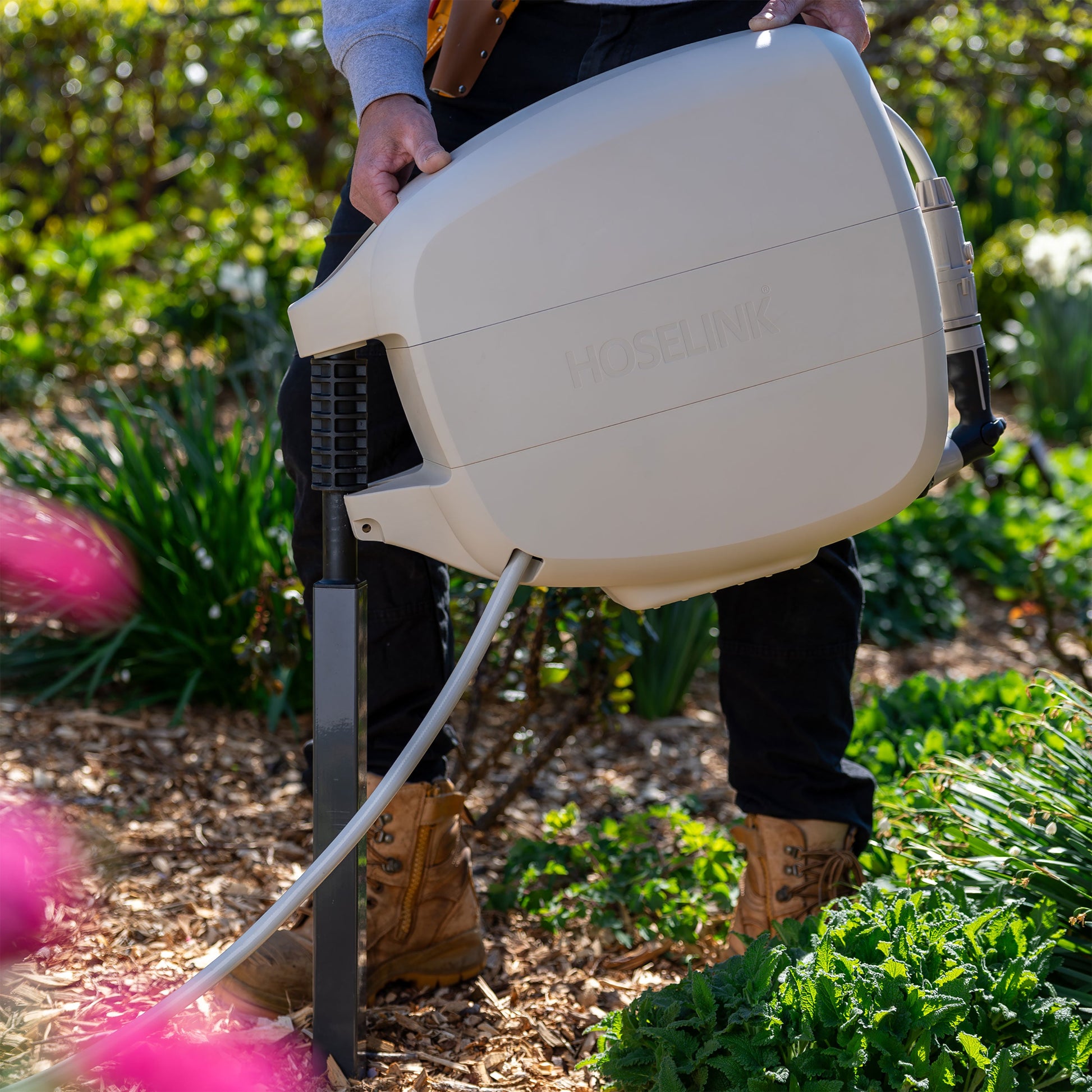 beige evolve retractable hose reel being mounted on an evolve mounting post is a mulch covered garden with green plants