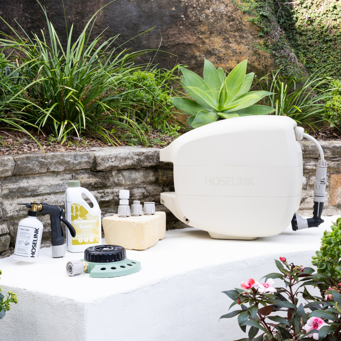 Evolve Beige Retractable Hose Reel sitting next to a Fertiliser Spray Mixer, Adapter Pack, 9-pattern Sprinkler and Seaweed Tonic on a white stone bench in front of a garden bed