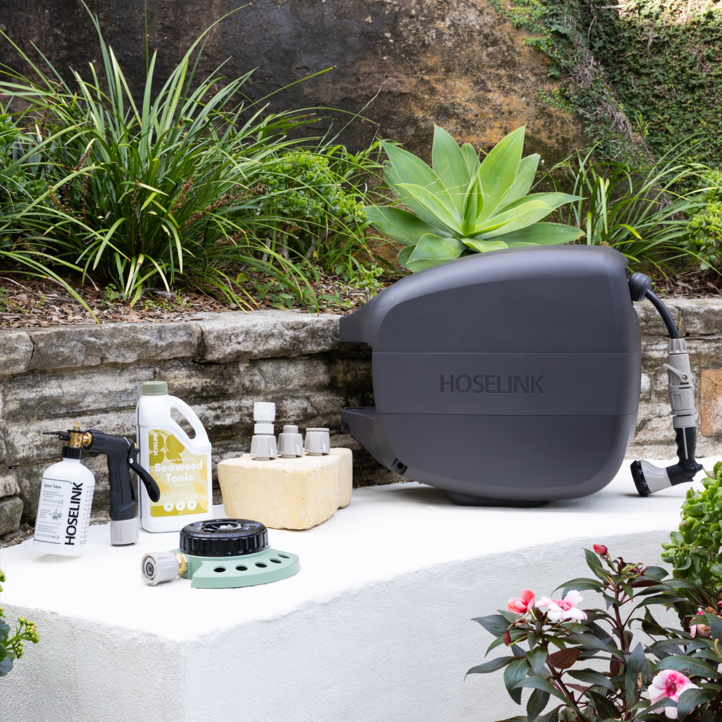 Charcoal Evolve Retractable Hose Reel sitting next to a Fertiliser Spray Mixer, Seaweed Tonic, Adapter Pack and 9-pattern Sprinkler on a white stone bench in front of a garden bed