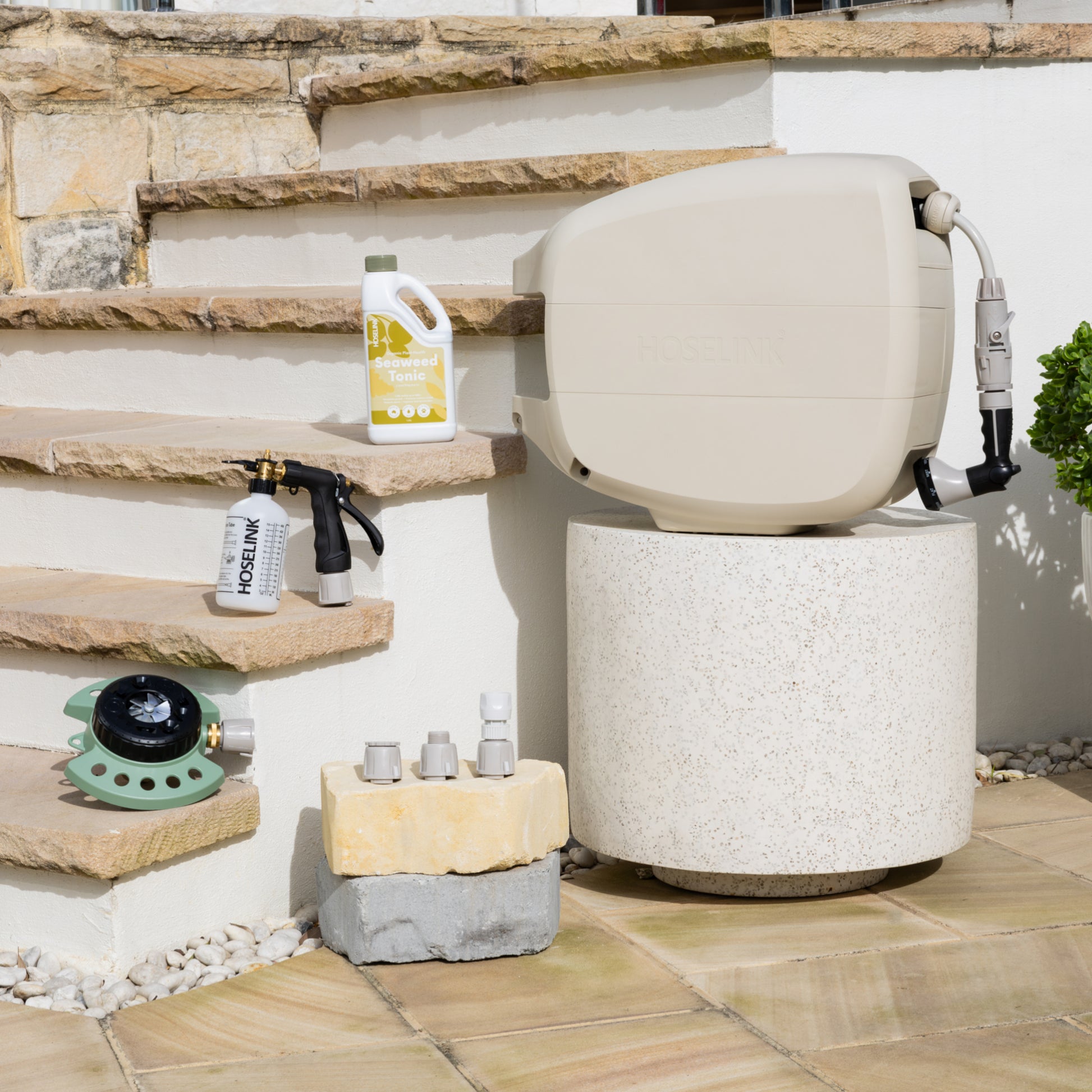 9-Pattern Sprinkler, Sea Weed Tonic Fertiliser Spray Mixer and Adapter Pack sitting on sandstone steps next to a white stone table with an Evolve Beige Retractable Hose Reel on it.